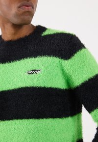 Maglione a righe nere e verdi realizzato in materiale peloso, con scollatura tonda e una piccola etichetta con logo nella parte anteriore.