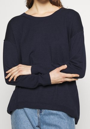 Kvinde iført en mørk marineblå langærmet trøje med rund hals, krydser armene, kombineret med lyseblå denimjeans.