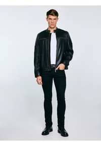 Fiatal férfi fekete bőrdzsekit, fehér inget, fekete nadrágot és fekete csizmát visel, egyszerű világos háttér előtt áll.