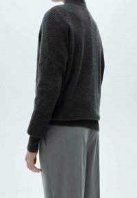 Personne portant un pull en maille texturée gris foncé et un pantalon ajusté gris clair, vue de dos sur un fond uni.