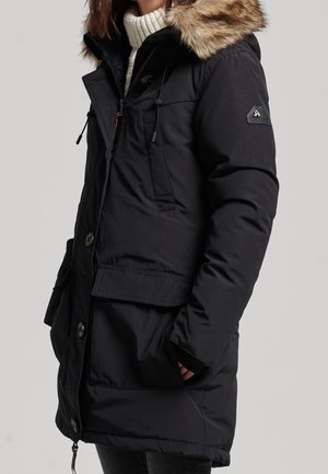 Black winter parka with fur-trimmed hood worn over a white turtleneck sweater, featuring button and zipper closure and front flap pockets.