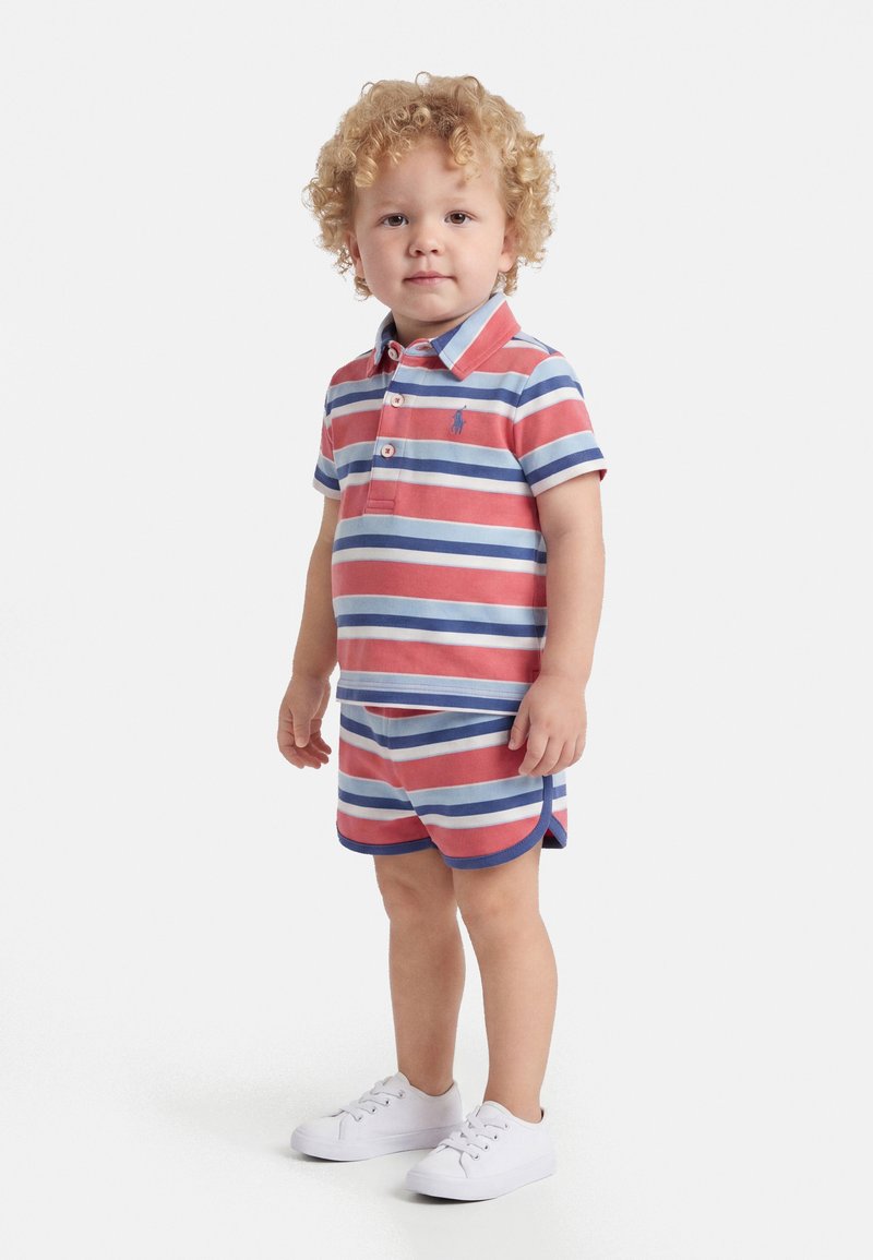 Jeune enfant aux cheveux bouclés portant un polo rayé rouge, blanc et bleu ainsi qu'un short assorti avec des baskets blanches, debout sur un fond blanc.