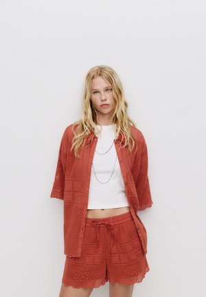 Chemise légère couleur rouille à manches courtes avec des boutons, assortie à un short texturé avec taille à cordon de serrage et ourlets en forme de vagues.