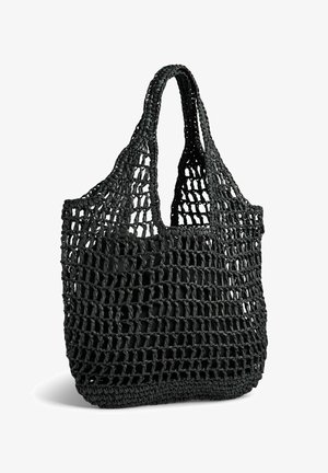Sac cabas noir en maille ouverte tissée avec de larges anses et un fond solide, debout sur fond blanc.