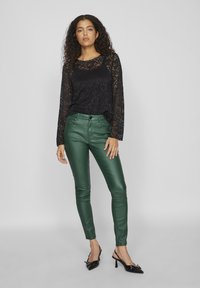 Black lace long-sleeve top, paired with fitted green leather pants and black pointed-toe heels, showcasing a sleek, modern look.