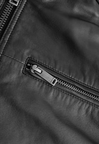 Jachetă din piele neagră cu un buzunar cu fermoar și accesorii din argint, textură netedă și detalii de cusătură subtile pe margini.