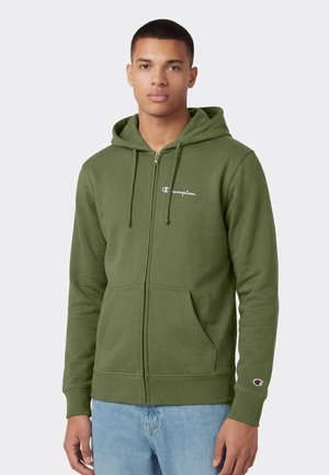 Jonge man draagt een olijfgroene hoodie met rits, voorzien van een capuchon en voorzakken, gecombineerd met lichtblauwe jeans, staand tegen een effen achtergrond.