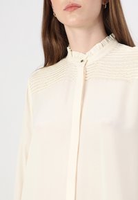 Cream-colored blouse featuring a ruffled collar and layered texture design on the shoulders, with a button closure at the front.