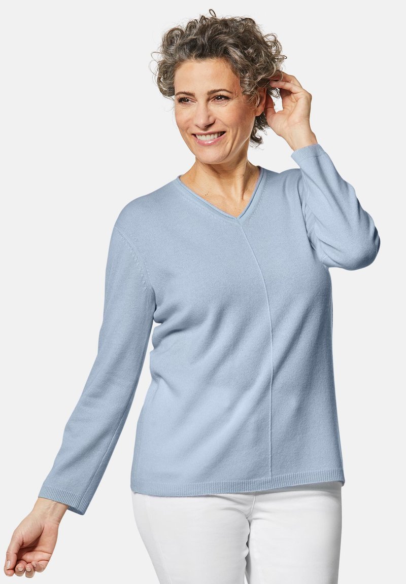 Helles, blaues, langärmliges Sweatshirt aus weichem Material mit V-Ausschnitt und subtilen diagonalen Nahtdetails. Kombiniert mit weißen Hosen.