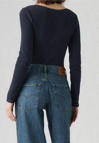 Person med marinblå räfflad långärmad topp och högmidjade blå denimjeans sedd bakifrån mot en enkel bakgrund.