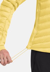 Tirando a mano il cordino regolabile dell'orlo della giacca, che mostra il tessuto trapuntato e la tasca con zip, indossata sopra pantaloni neri.