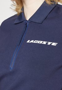 Gros plan d'une personne portant un polo bleu marine avec une fermeture éclair et un logo Lacoste blanc sur la poitrine.