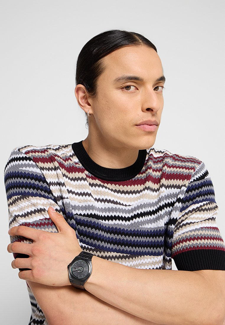 Joven con cabello largo y oscuro, vestido con un suéter colorido de patrón en zigzag, cruzando los brazos, lleva un reloj de pulsera negro y un pendiente de aro plateado.