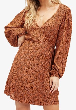 Kvinde i rust-orange slå-om-kjole med leopardprint, lange puffede ærmer og V-udskæring, accessoriseret med et guldhalskæde.