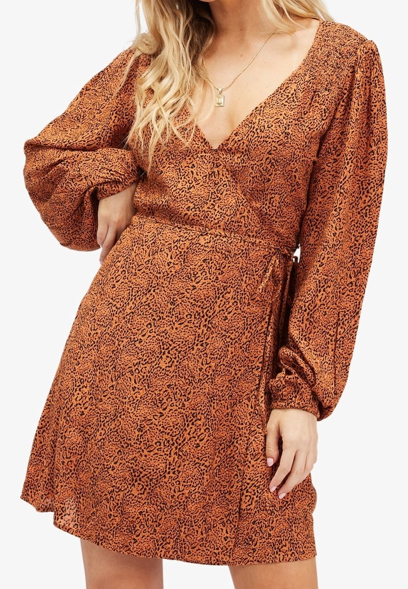 Femme portant une robe portefeuille à imprimé léopard rouille-orange avec de longues manches bouffantes et un décolleté en V, accessoirisée d'un collier en or.