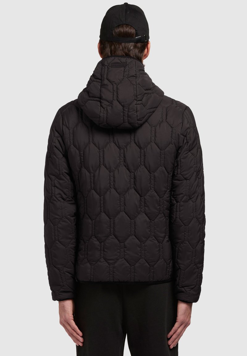 Doudoune noire avec un motif hexagonal, capuche et tissu texturé. Présente des coutures élastiques et une coupe courte, portée avec un pantalon sombre.