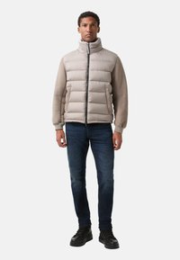 Lys brun polstret jakke med høj krave og sort lynlås, kombineret med mørkeblå jeans og sorte snørestøvler, stående på en hvid baggrund.