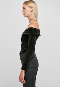 Svart kordfløyels off-the-shoulder topp med lange ermer, tettsittende design, glatt tekstur og sømdetaljer, kombinert med mørke jeans.
