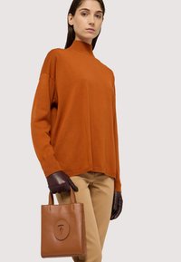 Brązowa skórzana torba na ramię z okrągłym logo, gładka tekstura. Modelka ma na sobie pomarańczowy sweter, brązowe rękawiczki i beżowe spodnie, prezentując luźny krój.