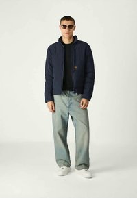 Homem de casaco acolchoado azul escuro, camisa preta, calças de ganga largas azul claro, tênis brancos e óculos de sol, de pé contra um fundo simples.