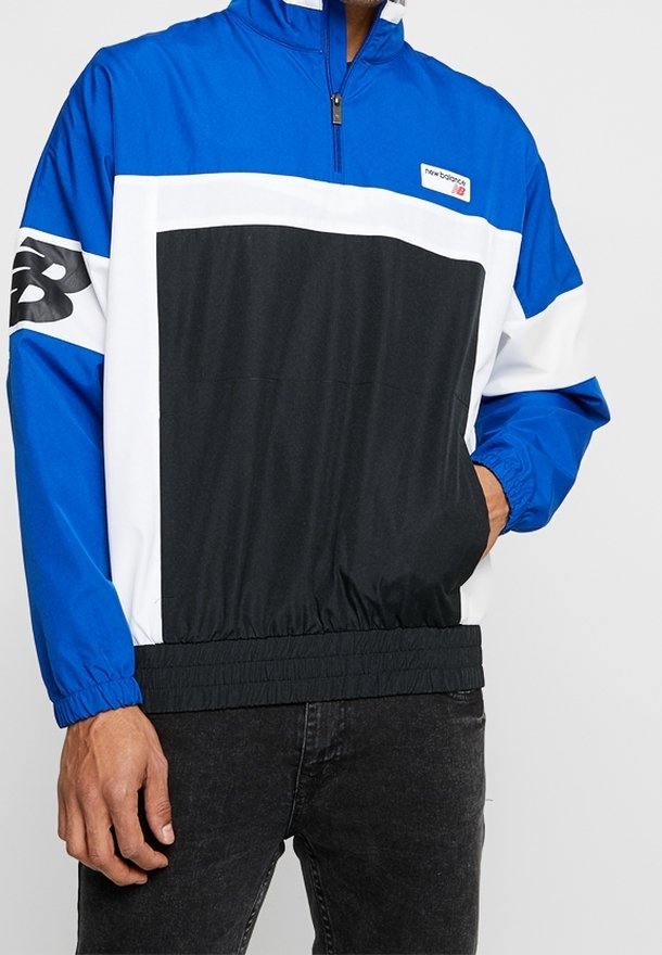 Lätt vindjacka i blått, svart och vitt. Halvdragkedja, elastiska ärmslut och nederkant, har en stor svart panel på framsidan och logotyp.