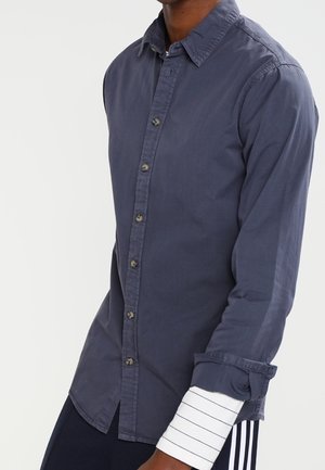 Homme portant une chemise gris foncé à boutons par-dessus une chemise à manches longues blanche avec de fines rayures noires, debout devant un fond uni.