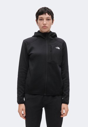 Persona con cabello corto y oscuro que lleva una sudadera con capucha negra con cremallera, bolsillo en el pecho y el logo de The North Face sobre un fondo claro.