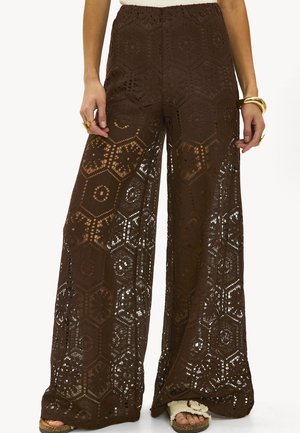 Pantalons en crochet à large jambe de couleur marron avec des motifs hexagonaux, portés avec des sandales blanches et des bijoux en or aux mains et poignets du mannequin.