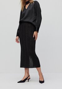 Blusa nera in satin con scollo a V e maniche lunghe abbinata a una gonna nera testurizzata di media lunghezza, caratterizzata da sottili pieghe e uno spacco laterale.
