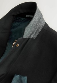 Veste noire texturée avec doublure intérieure de col grise, fermeture par boutons-pression dorés et tissu intérieur foncé à motifs visible.