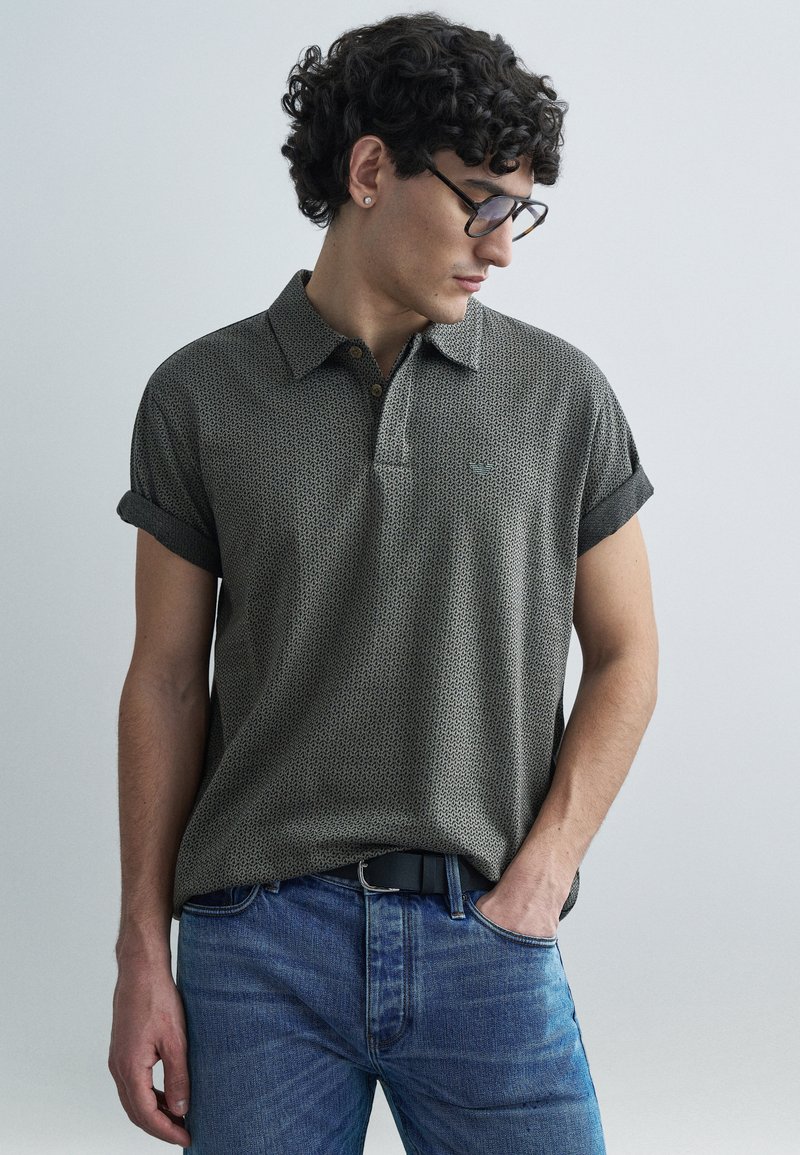 Polo à manches courtes en tissu vert foncé à motifs, avec un col et des revers de manche retroussés, porté avec un jean bleu et une ceinture noire.