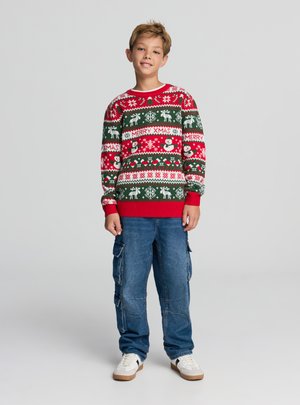 Maglione di Natale verde, rosso e bianco con decorazioni di renne, fiocchi di neve e pupazzo di neve; indossato con pantaloni cargo blu e sneaker bianche.