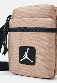 Borsa a tracolla in tessuto beige chiaro con superficie testurizzata. Presenta una cerniera nera e una toppa nera con il logo Jumpman bianco. Tracolla regolabile.