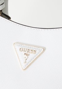 Borsa a mano bianca con texture e logo triangolare dorato che presenta "GUESS USA" e un punto interrogativo, insieme a bordi neri a contrasto.