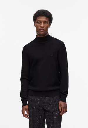 Black turtleneck sweater with long sleeves, textured knit, and subtle logo accent. Paired with fitted pants featuring a checked pattern.