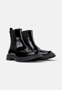 Botas negras hasta el tobillo con un acabado brillante, paneles laterales elásticos y una suela resistente, posicionadas sobre un fondo blanco.