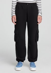 Czarne spodnie cargo wykonane z miękkiego materiału, z bocznymi kieszeniami, elastycznym paskiem w talii i elastycznymi mankietami przy kostkach. Połączone z niebieskimi trampkami.