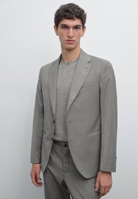 Hellgraue Anzugjacke mit tailliertem Schnitt, Revers mit Kerbe und zwei Fronttaschen, kombiniert mit einem strukturierten grauen Hemd darunter.