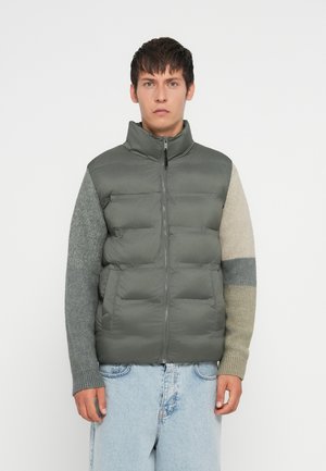 Hombre con chaleco de plumas verde y mangas de punto multicolores, usando jeans de mezclilla de color azul claro, de pie contra un fondo blanco.