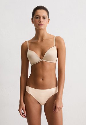 Woman standing against white wall wearing beige bra and underwear set, facing forward with a neutral expression and hands relaxed by sides.