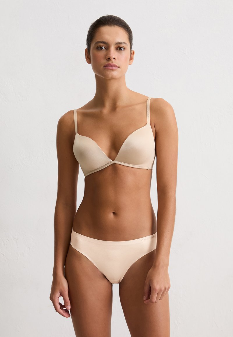 Vrouw die tegen een witte muur staat met een beige bh en ondergoed set, recht vooruit kijkend met een neutrale uitdrukking en handen ontspannen naast zich.