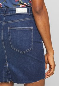 Džínová mini sukně v tmavě modré barvě s roztrhaným lemem, se dvěma zadními kapsami a viditelnou nášivkou značky na pase.