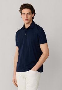 Hombre con camisa polo de manga corta azul marino y pantalones blancos, de pie con una mano en el bolsillo contra un fondo gris liso.