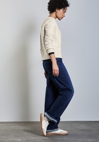 Cremfarbener Pullover mit einem strukturierten Muster, kombiniert mit dunkelblauen Jeans und weißen Sneakers. Die Jeans sind an den Knöcheln hochgekrempelt.
