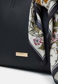 Bolso de mano de cuero negro con una bufanda de seda de patrón floral atada al asa. Detalle de placa de la marca en dorado en la parte frontal. Textura suave.