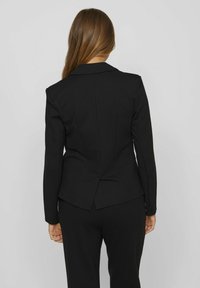 Blazer nero su misura con una silhouette aderente, spalle imbottite, revers a punta e spacco sul retro. Realizzato in un tessuto liscio e strutturato.