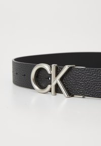 Cinturón de cuero negro con acabado texturizado, que presenta una hebilla circular plateada con las letras "CK". Ajustable con pequeños agujeros para un mejor calce.
