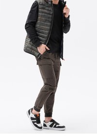 Gepolsterte olivgrüne Weste über einem schwarzen Langarmshirt, kombiniert mit braunen Cargo-Hosen mit Taschen und "STATE"-Druck in Kniehöhe. Schwarz-weiße Sneakers mit orangefarbenen Akzenten.