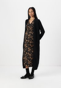 Cardigan lungo nero sopra un vestito nero con un motivo floreale marrone, abbinato a calze nere e scarpe nere lucide.
