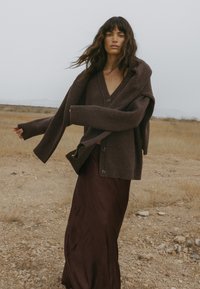 Rudas megztas kardiganas su V formos iškirpte, per didelis, su mygtukais, uždėtas ant tamsiai burgundiško satino sijono, natūralioje aplinkoje.